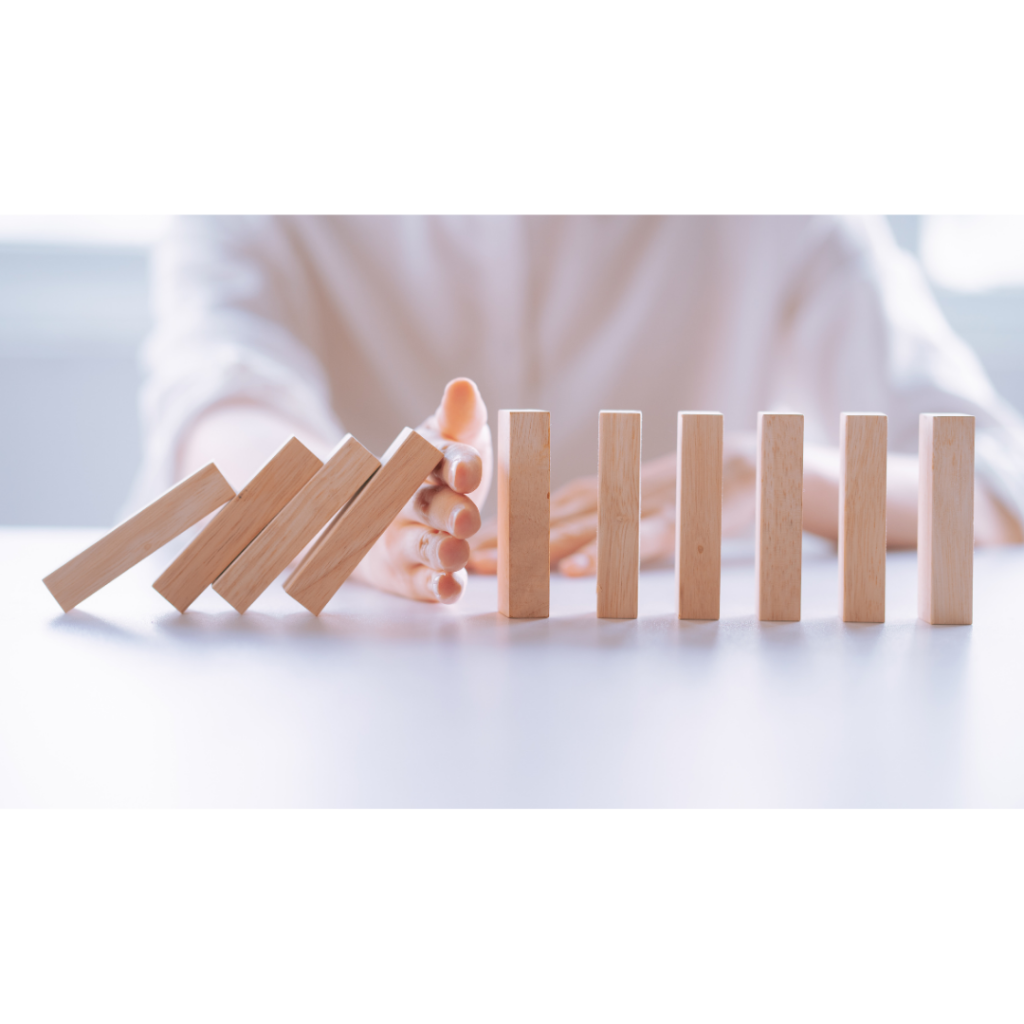 The image depicts a close-up of a hand stopping a line of falling dominoes just before the chain reaction continues. This powerful visual symbolizes proactive crisis management, where timely intervention prevents a small issue from escalating into a larger disaster. The hand represents leadership or a crisis management team, stepping in to halt the spread of chaos and maintain control, emphasising the importance of quick, decisive action in preventing further damage.