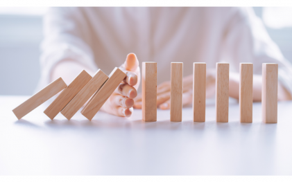 The image depicts a close-up of a hand stopping a line of falling dominoes just before the chain reaction continues. This powerful visual symbolizes proactive crisis management, where timely intervention prevents a small issue from escalating into a larger disaster. The hand represents leadership or a crisis management team, stepping in to halt the spread of chaos and maintain control, emphasising the importance of quick, decisive action in preventing further damage.